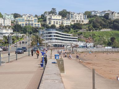 A beachfront promenade with people walking and cycling near a sandy beach and buildings on a hillside at Apartment 17 Abbey Sands in Torquay