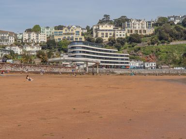 A sandy beach with people sitting and walking near a modern curved building and hillside houses at Apartment 17 Abbey Sands in Torquay