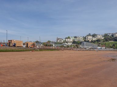 A sandy beach with people sitting near wooden huts and houses on a hillside in the background at Apartment 17 Abbey Sands in Torquay