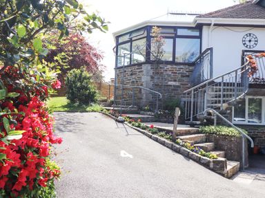 Driveway with flowers along the edges and stone steps leading to a house with large windows and a clock on the wall at Lake View Swanpool Nr Falmouth