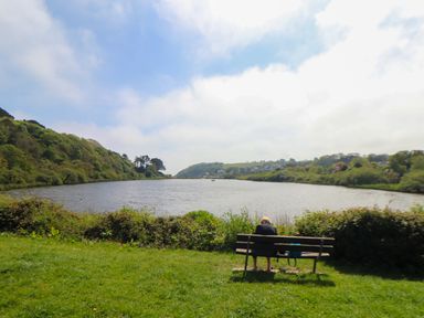 A person sitting on a bench by a lake with hills and houses in the background at Lake View in Swanpool Nr Falmouth