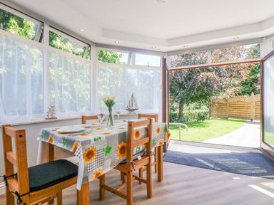 A dining area with a wooden table and chairs covered with a sunflower tablecloth next to large windows and open door to a garden at Lake View in Swanpool Nr Falmouth