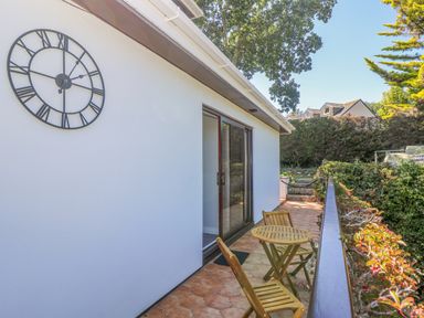 An outdoor patio area with a small wooden table and two chairs adjacent to a white wall with a large clock and sliding glass door at Lake View in Swanpool Nr Falmouth