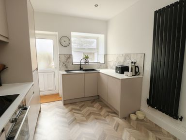 A kitchen with beige cabinets a black sink a toaster kettle and cups on the counter and a black vertical radiator at 1 Mona Terrace in Llanfachraeth