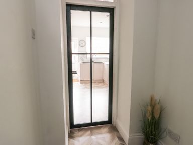 A hallway with a glass door leading to a kitchen with a clock on the wall and a potted plant in the corner at 1 Mona Terrace in Llanfachraeth