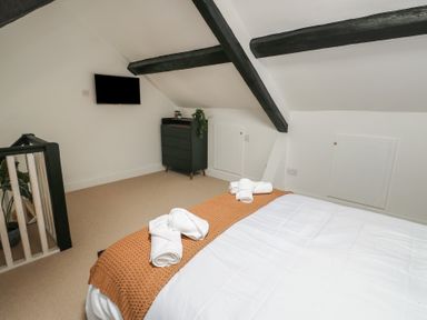 A bedroom with a bed covered in white and orange blankets a black dresser a wall mounted television and exposed ceiling beams at 1 Mona Terrace in Llanfachraeth