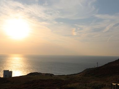 A coastal scene with the sun setting over the ocean a lighthouse on the right and a building on the left at 1 Mona Terrace in Llanfachraeth