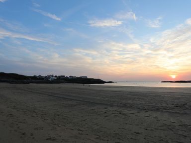 A sandy beach with houses in the distance and the sun setting over the sea at 1 Mona Terrace Llanfachraeth
