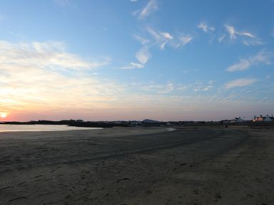 A sandy beach near a coastal village with houses and a setting sun at 1 Mona Terrace in Llanfachraeth