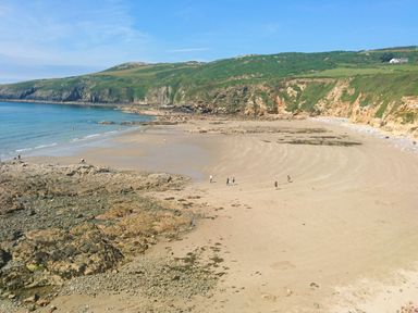 A sandy beach with scattered rocks and people walking near cliffs and green hills at 1 Mona Terrace in Llanfachraeth