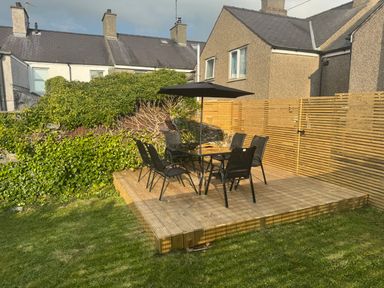 A wooden deck with a table umbrella and six chairs in a garden surrounded by greenery and houses at 1 Mona Terrace in Llanfachraeth