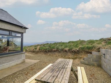 An outdoor area with a wooden picnic table next to a house with large windows and grassy hillside at Castell Y Garn near Newport Beach Pembrokeshire