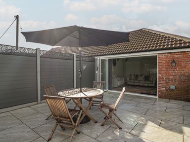 An outdoor patio with a round wooden table and four chairs under a large umbrella next to a house with sliding glass doors at Doubletree Cottage in Swainsford near Mere