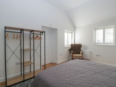 A bedroom with a grey quilted bedspread an open wardrobe with hangers a chair with a fur throw and two windows with blinds at Singletree Cottage in Swainsford near Mere