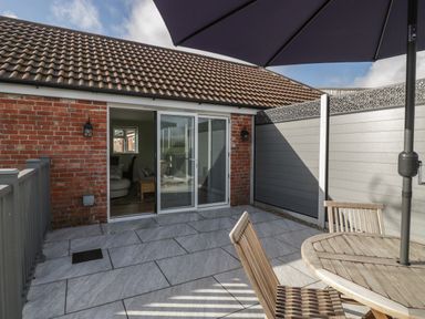 An outdoor patio area with wooden chairs and table under a large umbrella next to a brick building with sliding glass doors at Singletree Cottage in Swainsford near Mere