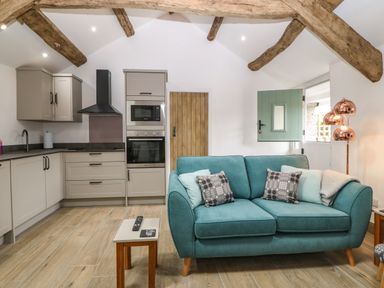 A kitchen and living area with wooden beams on the ceiling a teal sofa with cushions a small table and built in kitchen units at Y Stabl in Dyffryn Ardudwy