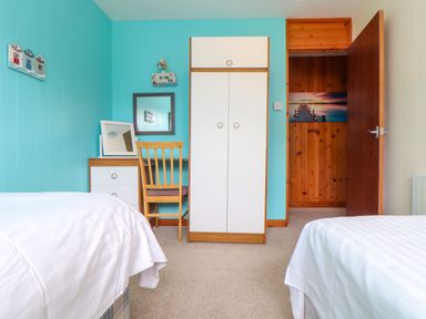 A bedroom with two beds a wooden wardrobe a desk with a chair and mirror and a door leading to a wooden-paneled hallway at Skylarks Trevellas Nr St Agnes