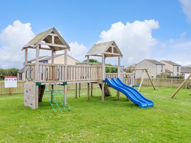A wooden playground structure with two blue slides swings and climbing features on grass at Skylarks Trevellas Nr St Agnes