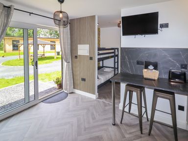 A room with bunk beds a wall-mounted television a counter with two stools and sliding glass doors overlooking a grassy area at Valley Lodge 4 - North Wales Eco Lodges in Graigfechan near Ruthin