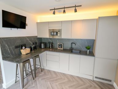 A kitchen with grey cabinets a black countertop a mounted TV and two metal stools at Valley Lodge 4 - North Wales Eco Lodges in Graigfechan near Ruthin