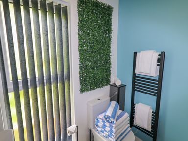 A bathroom with vertical blinds a toilet with stacked blue and white towels a towel rack with white towels and a green wall decoration at Valley Lodge 4 - North Wales Eco Lodges in Graigfechan near Ruthin