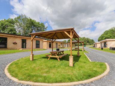 Picnic tables with wooden shelters on grass between gravel pathways with wooden lodges in the background at Valley Lodge 4 - North Wales Eco Lodges in Graigfechan near Ruthin