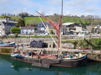 A black sailing boat docked in a harbor with houses and green hills in the background at 20 Keay Heights in St Austell