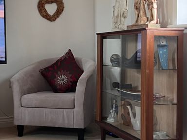 A beige armchair with a patterned cushion next to a wooden glass display cabinet with various decorative items at 20 Keay Heights in St Austell