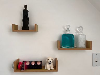 Wooden wall shelves with a black statue, two glass bottles, a box with bath bombs, and a small white stuffed dog at 20 Keay Heights in St Austell