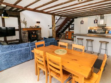 An open plan room with a wooden dining table and chairs a blue sofa a stone fireplace a TV and a kitchen area with bar stools and pendant lights at The Old Smithy in Lynton