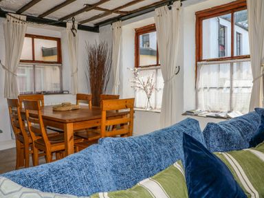 A dining area with a wooden table and chairs next to windows with curtains and a blue sofa at The Old Smithy in Lynton