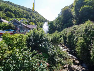 A river flowing through greenery with bushes and trees near a stone building and a flagpole at The Old Smithy in Lynton