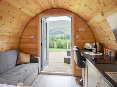 A small wooden cabin interior with a gray sofa, kitchenette with bar stools, and open double doors leading to a deck with a view of green hills at Caban Tirion in Dinas Mawddwy near Dolgellau