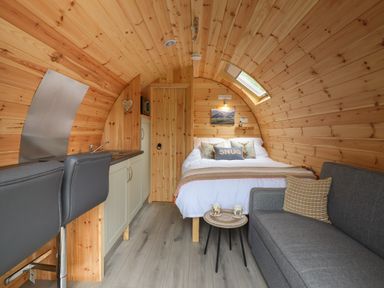 A small wooden cabin interior with a bed, kitchenette, two bar chairs, a grey sofa, and a small round table with mugs at Caban Tirion in Dinas Mawddwy near Dolgellau