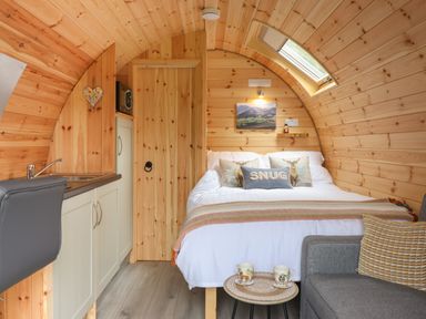 A wooden pod interior with a bed, pillows, small kitchen area, and a chair at Caban Tirion in Dinas Mawddwy near Dolgellau