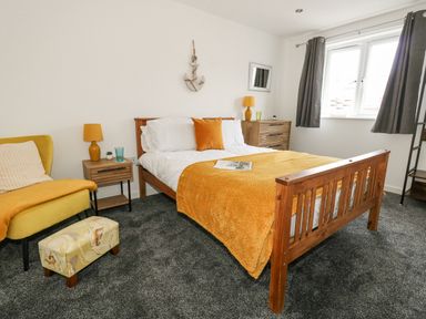 A bedroom with a wooden bed and orange bedding a yellow armchair with a footrest brown bedside tables with lamps and a window with curtains at Dotties Retreat in Weymouth