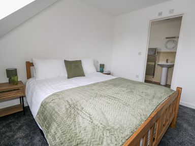 A bedroom with a wooden bed, green bedding, two nightstands with lamps and glasses, and an open door showing a bathroom at Dotties Retreat in Weymouth