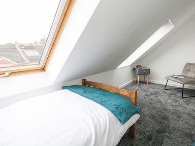 A bedroom with a single wooden bed a teal blanket a gray chair and a small gray bedside table under slanted windows at Dotties Retreat in Weymouth