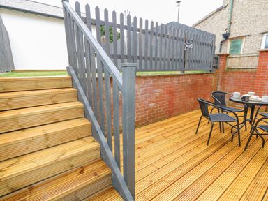 A wooden deck patio with black chairs and a table next to a brick wall and wooden stairs with a gray fence at Dotties Retreat in Weymouth