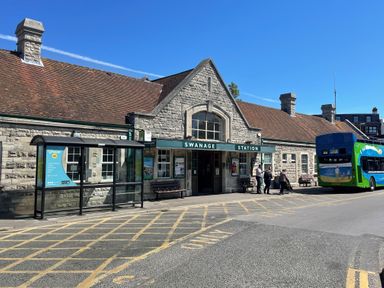 An exterior view of Swanage Station with a stone building a bus shelter and a bus with people standing nearby at The Swanage Rail Lookout in Swanage