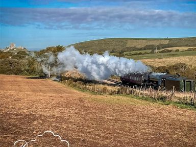 A steam train moving through a rural landscape with fields and hills at The Swanage Rail Lookout in Swanage