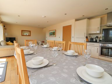 A dining table set with white bowls, plates, and wine glasses in a room with wooden chairs, a kitchen, and a living area at The Willows in Winnard's Perch near St Columb Major