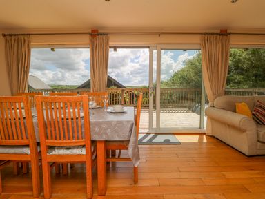 A dining area with a wooden table and chairs next to large glass sliding doors and beige sofas at The Willows in Winnard's Perch near St Columb Major