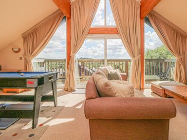 A living room with a sofa pillows pool table and large windows overlooking a balcony and trees at The Willows in Winnard's Perch near St Columb Major