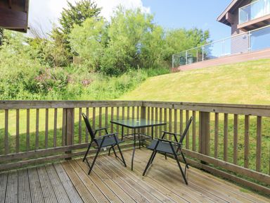 A wooden deck with two black chairs and a glass table overlooking a grassy yard with trees at The Willows Winnards Perch near St Columb Major