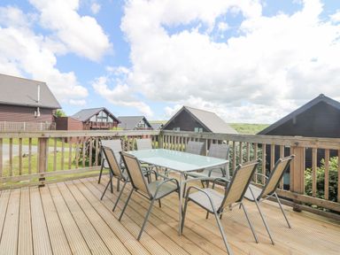 An outdoor wooden deck with a glass table and eight chairs overlooking nearby cabins at The Willows in Winnard's Perch near St Columb Major
