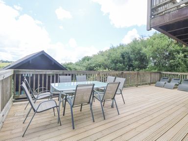 An outdoor wooden deck with a glass table surrounded by six metal chairs and three black lounge chairs at The Willows in Winnard's Perch near St Columb Major