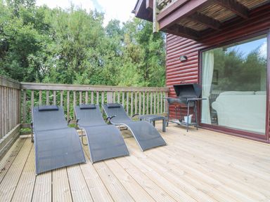 An outdoor wooden deck with three lounge chairs a small table a barbecue grill and glass door to interior at The Willows in Winnard's Perch near St Columb Major