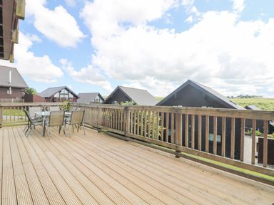 A wooden deck with a table and chairs overlooking dark wooden houses and green fields at The Willows in Winnard's Perch near St Columb Major