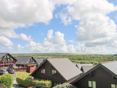 A group of wooden cabins with peaked roofs near a green field under a blue sky with clouds at The Willows Winnard's Perch near St Columb Major